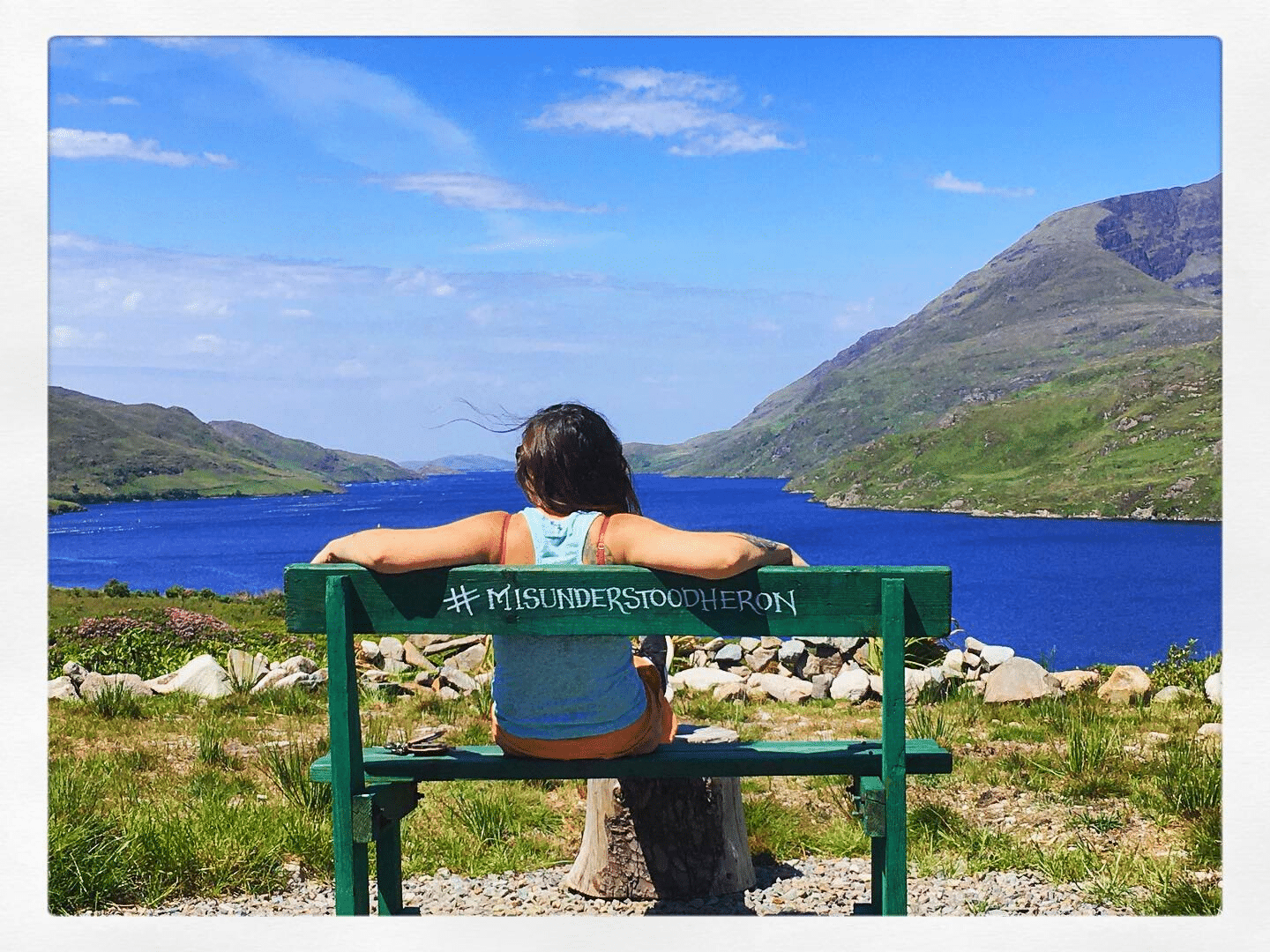 a woman sittting on a green bench facing a fjord with mountains on either side