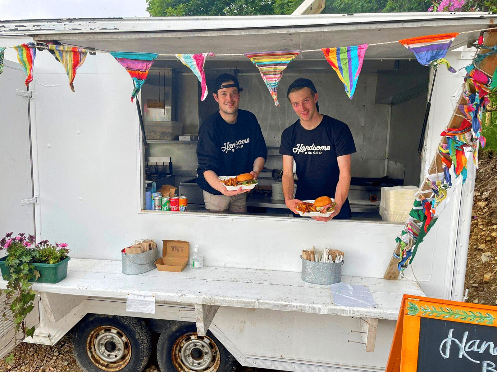 two men in a white food truck holding a plate of burger and chips